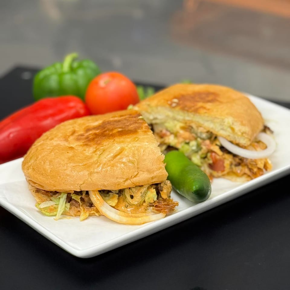 Two halves of a Mexican torta sandwich with meat, lettuce, and onions, served with a jalapeño.