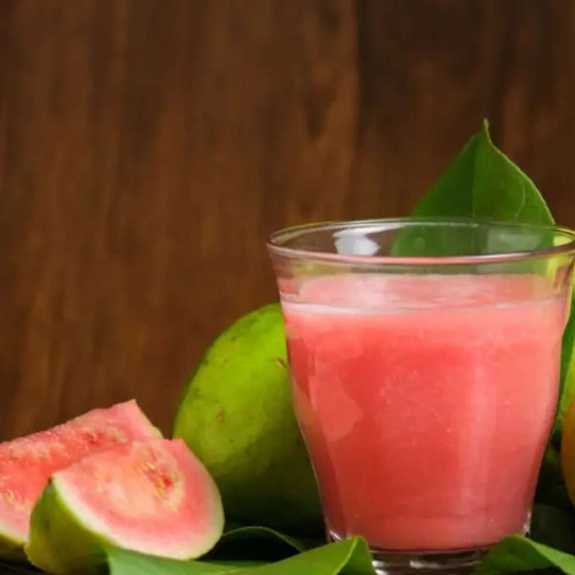 A glass of pink guava juice with fresh guava slices and leaves on a wooden background.