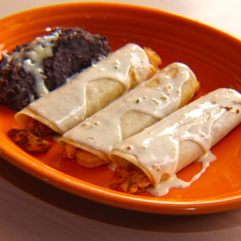 Three enchiladas covered in white sauce with a side of black beans on an orange plate.